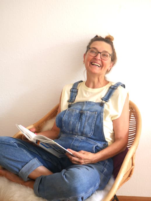 Woman sitting on a rattan chair holding a book and laughing out loud.