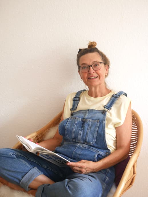 Woman sitting on a rattan chair holding a book and looking up to the camera.
