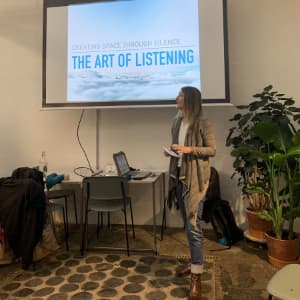 A woman standing in front of a big screen which shows a slide with The Art of Listening written on it.