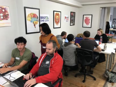 Two tables with people working on laptops, some in a group, some alone. One woman is standing.