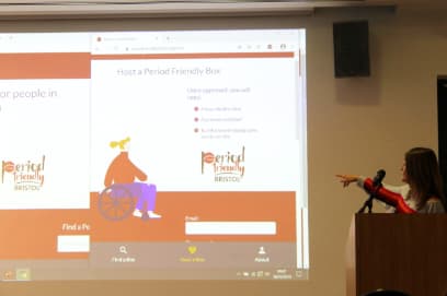A woman standing in front of a big screen, pointing to the display and talking into a microphone.